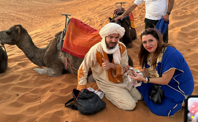 Un hombre y una mujer se sientan junto a un camello en el desierto al atardecer, vestidos con atuendos tradicionales. Otro hombre está cerca, acariciando un camello. El ambiente es cálido y sereno.