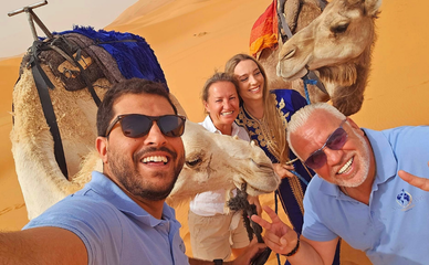 Un grupo de personas y dromedarios posan alegremente en el desierto, con amplias sonrisas y ropa informal. Las dunas de arena iluminan el soleado fondo.