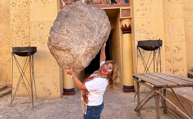A person playfully poses as if lifting a large rock in front of an Egyptian-themed building with hieroglyphics and vibrant decorations, exuding a fun, lighthearted vibe.