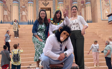 Cuatro personas posan en una escalera frente a un antiguo templo de estilo egipcio con coloridas columnas y jeroglíficos. Una de ellas lleva una máscara de mono.