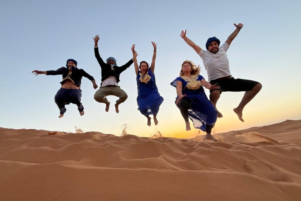 Cinco personas saltan en el aire sobre una duna de arena al atardecer, vestidas con ropa azul y blanca y pañuelos en la cabeza, con un cielo despejado de fondo.