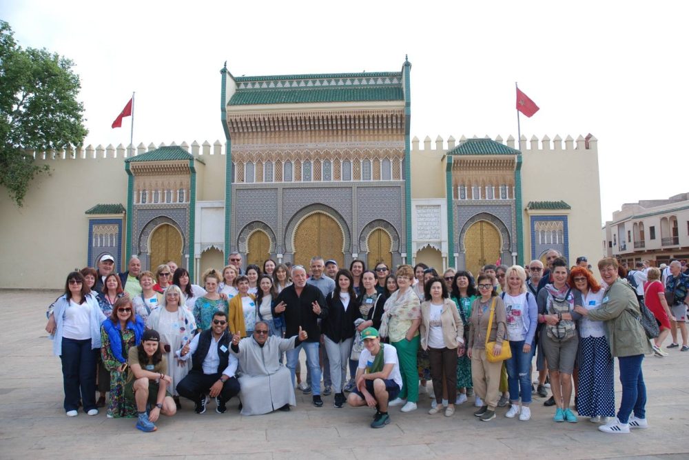 Un grupo diverso de personas posa sonriente frente a un edificio ornamentado con puertas arqueadas doradas y patrones intrincados, lo que sugiere una atmósfera animada y alegre.