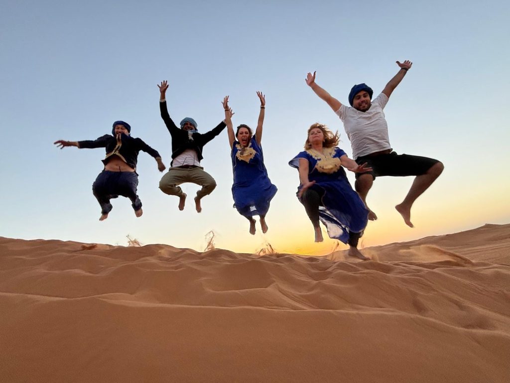 Cinco personas saltan en el aire sobre una duna de arena al atardecer, vestidas con ropa azul y blanca y pañuelos en la cabeza, con un cielo despejado de fondo.