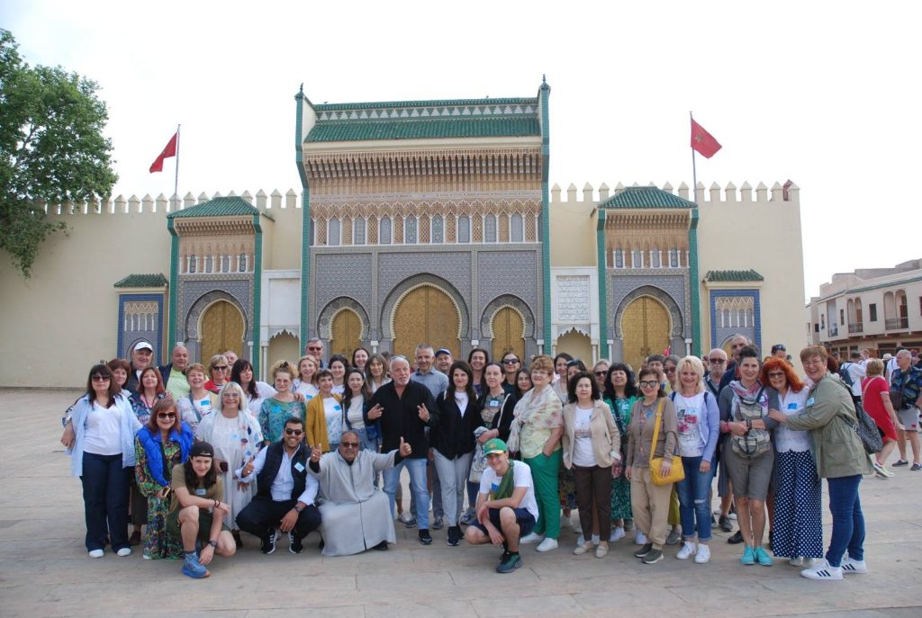 Un grupo diverso de personas posa sonriente frente a un edificio ornamentado con puertas arqueadas doradas y patrones intrincados, lo que sugiere una atmósfera animada y alegre.
