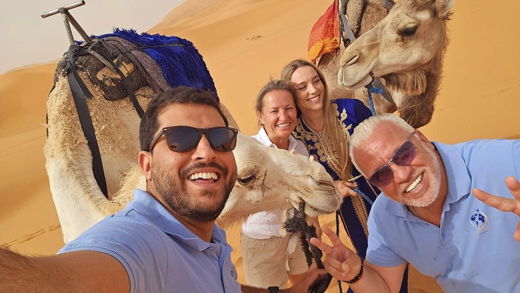 Un grupo de personas y dromedarios posan alegremente en el desierto, con amplias sonrisas y ropa informal. Las dunas de arena iluminan el soleado fondo.
